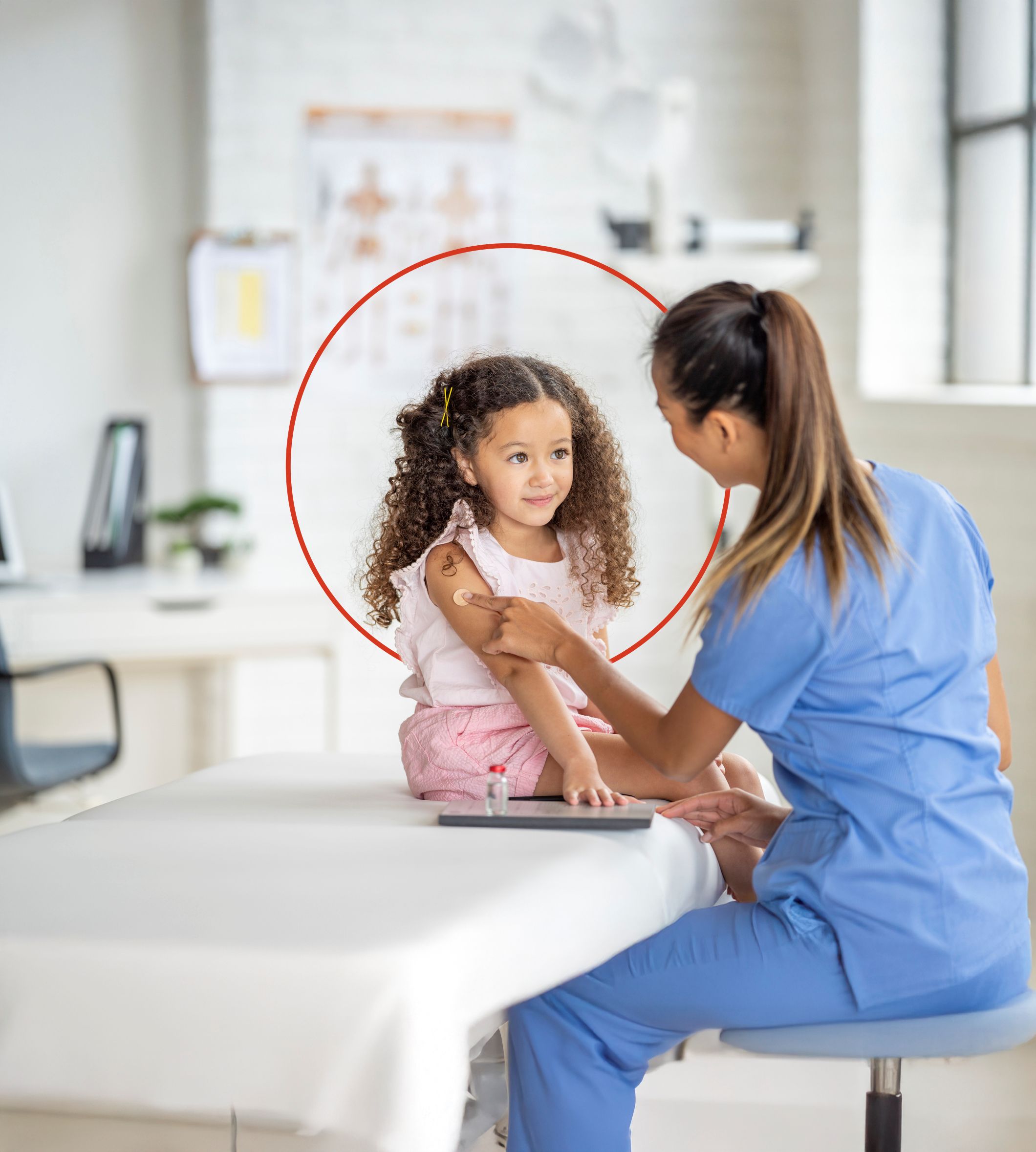 A sweet little girl of Latin decent, sits up straight on an exam table as her nurse places a bandage on her arm after giving her a vaccination. The nurse is wearing blue scrubs and smiling at the young girl to put her at ease.