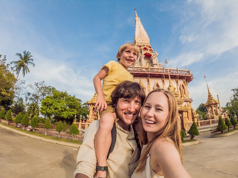 Familie mit Vater, Mutter und Kind (auf Papas Schultern) beim Sightseeing vor einem Tempel, im Hintergrund Palmen bei Sonnenschein.