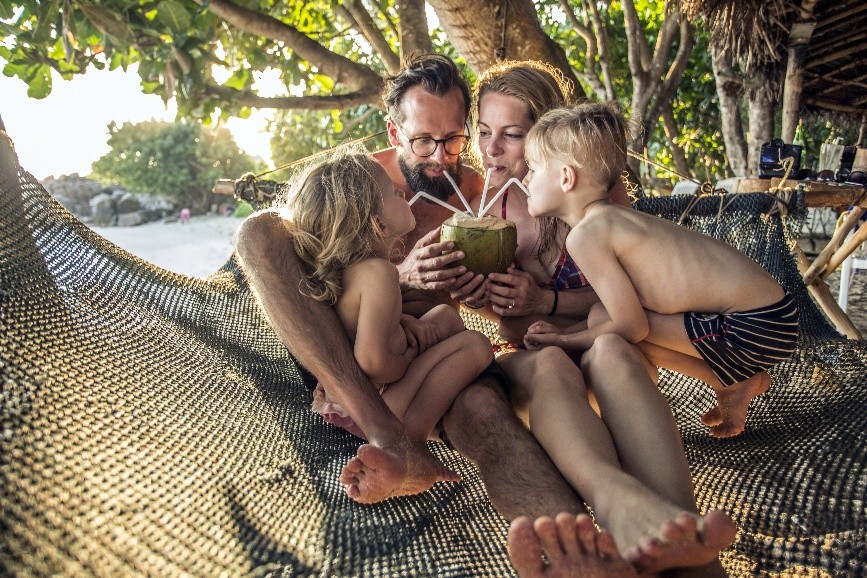 Familie mit Mutter, Vater, Junge und Mädchen sitzt am Strand und trinkt gemeinsam aus einer Kokosnuss, jeder mit eigenem Strohhalm.